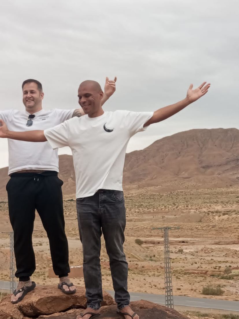 Two travellers with arms flung wide on a rock above a dry desert valley