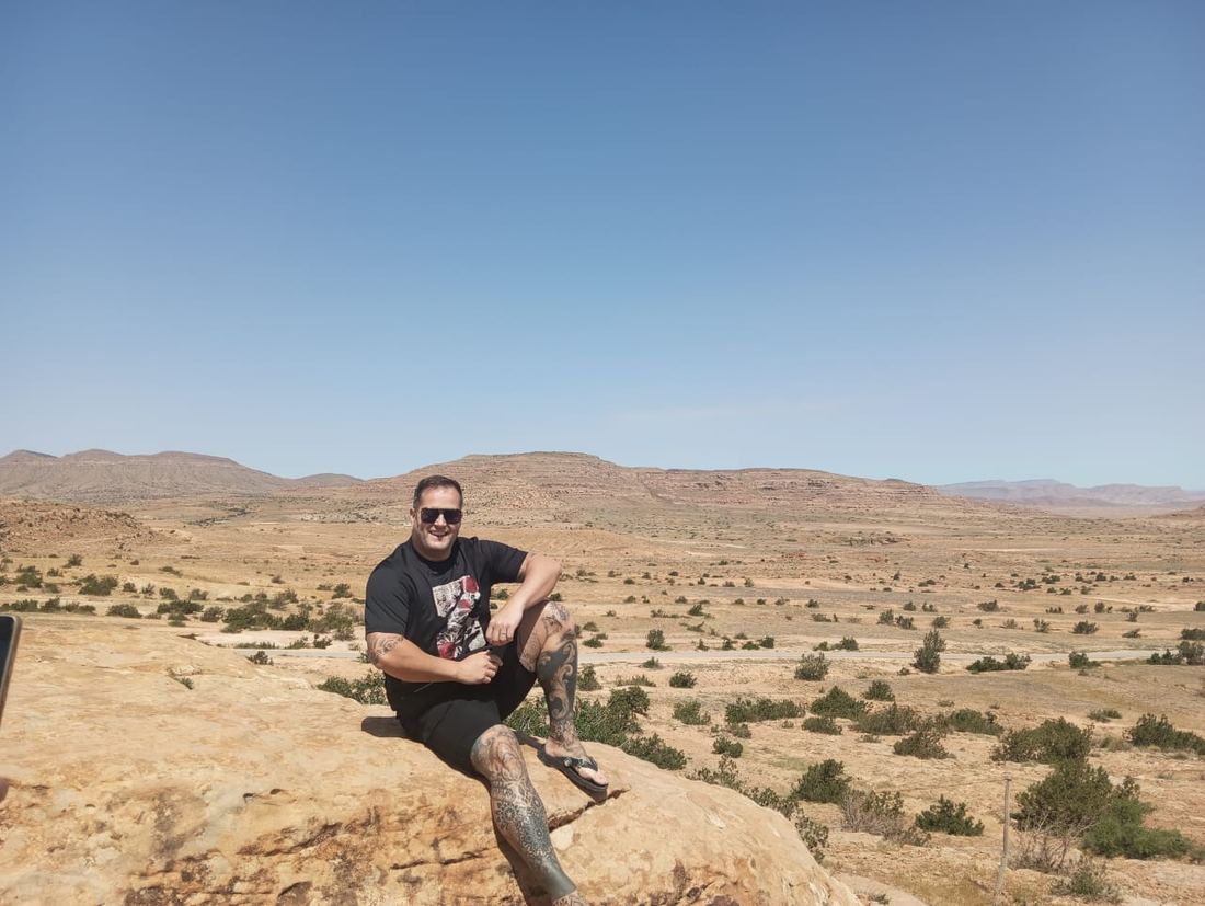A traveller sitting on a sandstone ledge above a wide desert plateau of scrub and distant escarpments