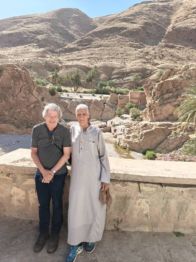 A traveller and a local host in a white gandoura on a wall above a palm-filled gorge