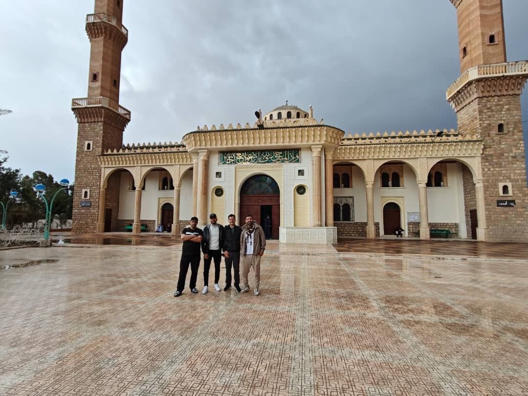 A group of travellers on the wide paved forecourt of a mosque with twin minarets