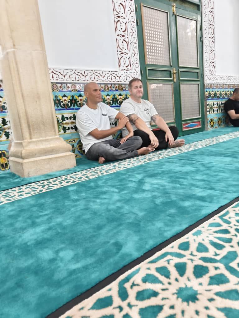 Two travellers sitting on the teal carpet of a mosque beside tiled walls