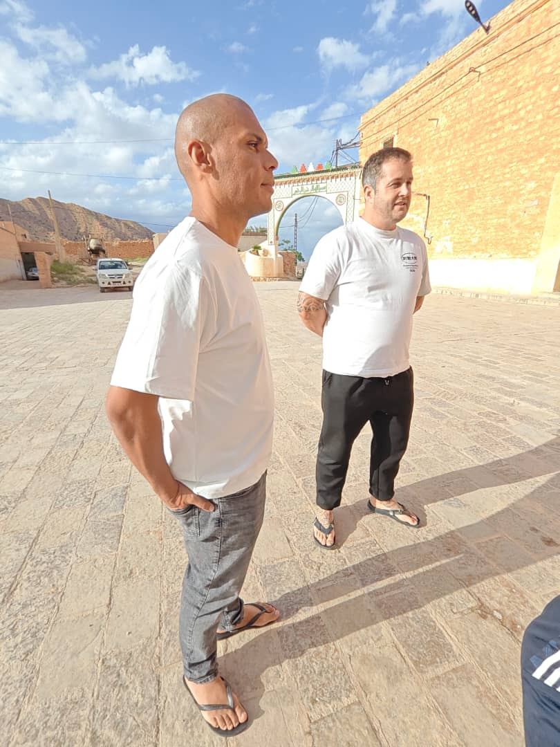 Two travellers on a paved village square in front of a painted ceremonial arch