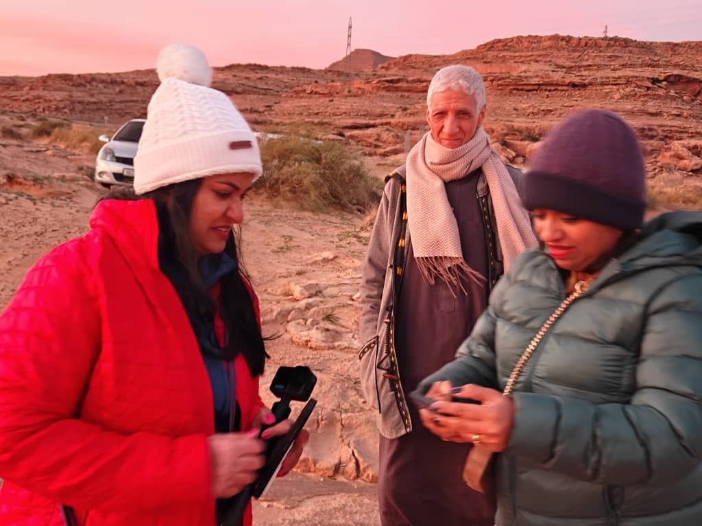 Two travellers and a local elder looking at a phone in the desert at dusk