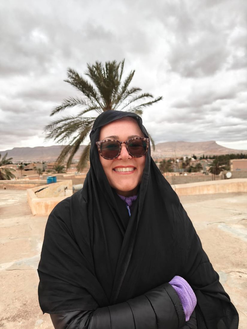 A smiling traveller wrapped in a black shawl on a rooftop, a palm tree behind