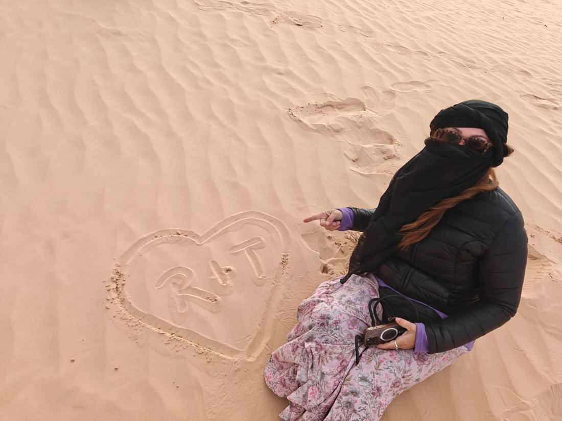 A traveller, face wrapped against the wind, beside initials drawn in a heart on the sand