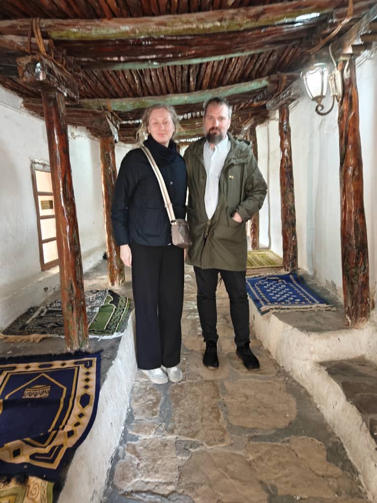 A couple standing in an old prayer hall roofed with palm trunks, prayer rugs laid on the stone floor