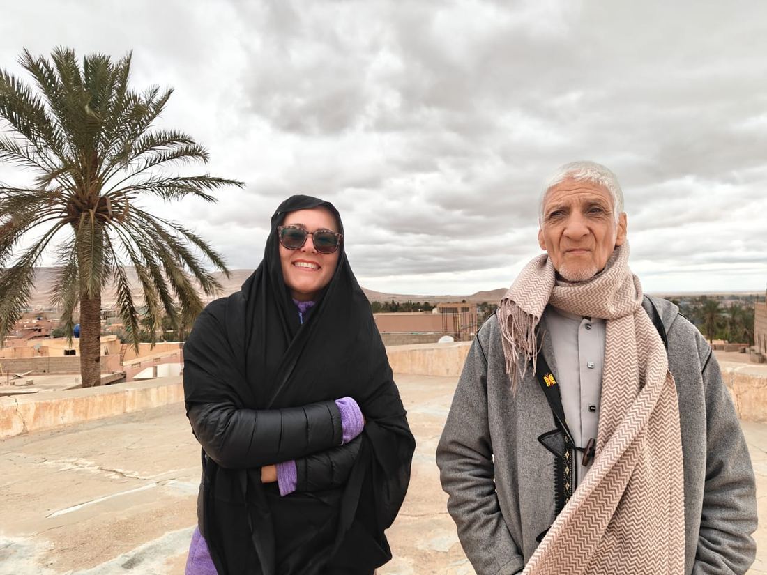 A traveller in a black shawl and a local elder on a rooftop terrace, palms and town beyond