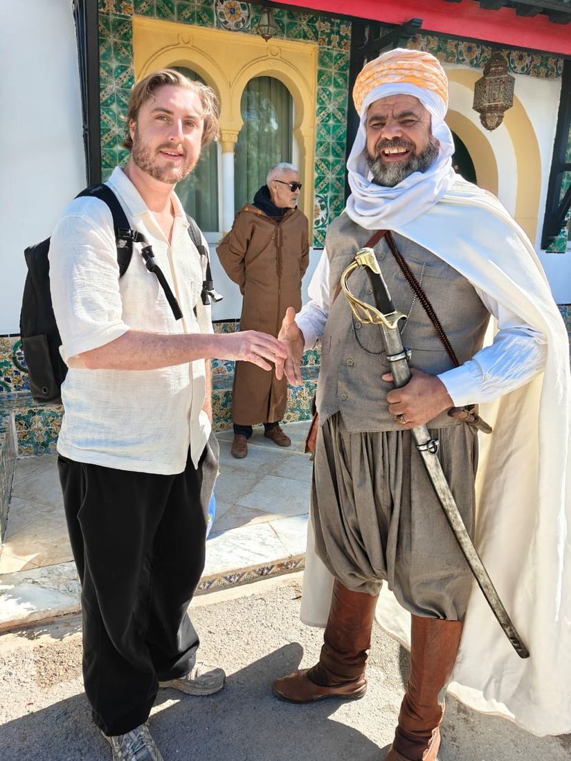 A traveller shaking hands with a man in traditional horseman's dress, a sabre at his side