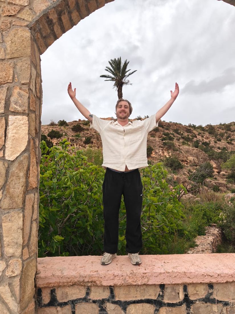 A traveller with arms raised under a stone arch above green hills