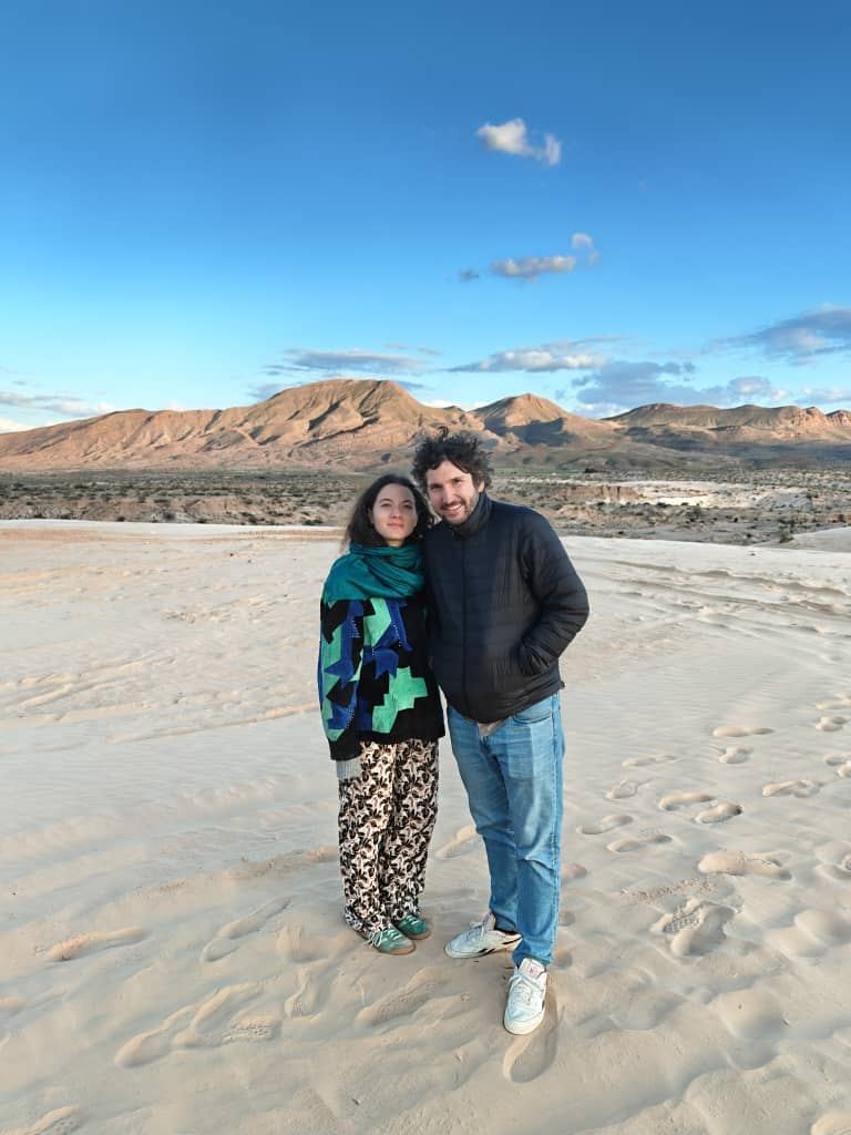 A couple standing on pale sand with desert mesas behind them late in the day