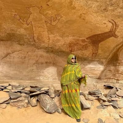 A woman in a green wrap beneath prehistoric paintings of a figure and an antelope in a Tassili rock shelter