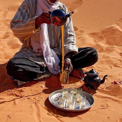 A Tuareg man in a white turban pouring tea from a height on the dunes