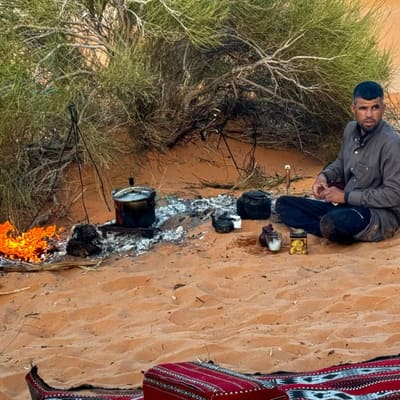 A Saharan guide brewing tea over an open fire in the sand, kettle and pots beside a patterned rug