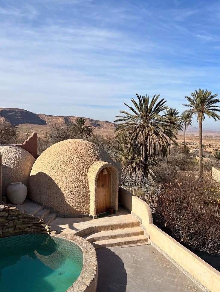 Domed earthen guest lodgings and a small pool at Bou Saâda, palms and desert hills beyond