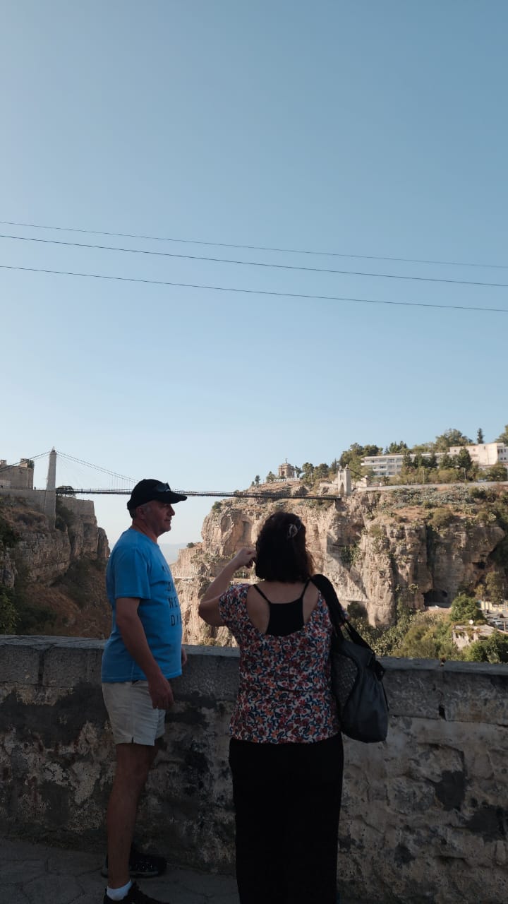 Two travellers at a viewpoint above the Rhumel gorge, the Sidi M'Cid suspension bridge strung across it behind them