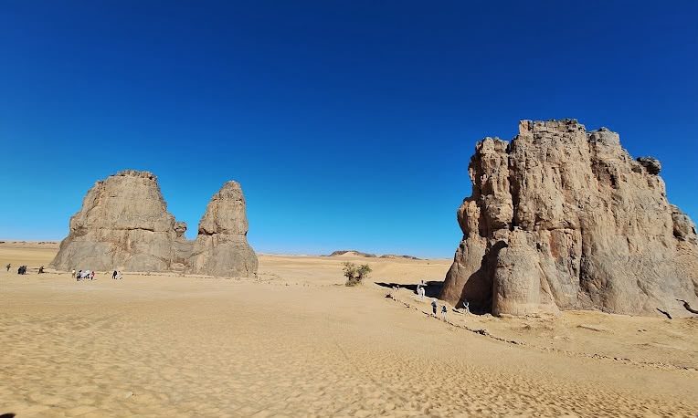 Two great rock massifs on an open sand plain, walkers at their foot giving the scale