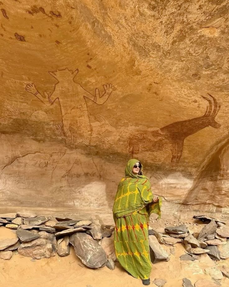 A woman in a green wrap beneath prehistoric paintings of a figure and an antelope in a Tassili rock shelter