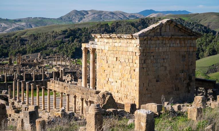A small Roman temple beside a colonnade at Djémila, mountains beyond