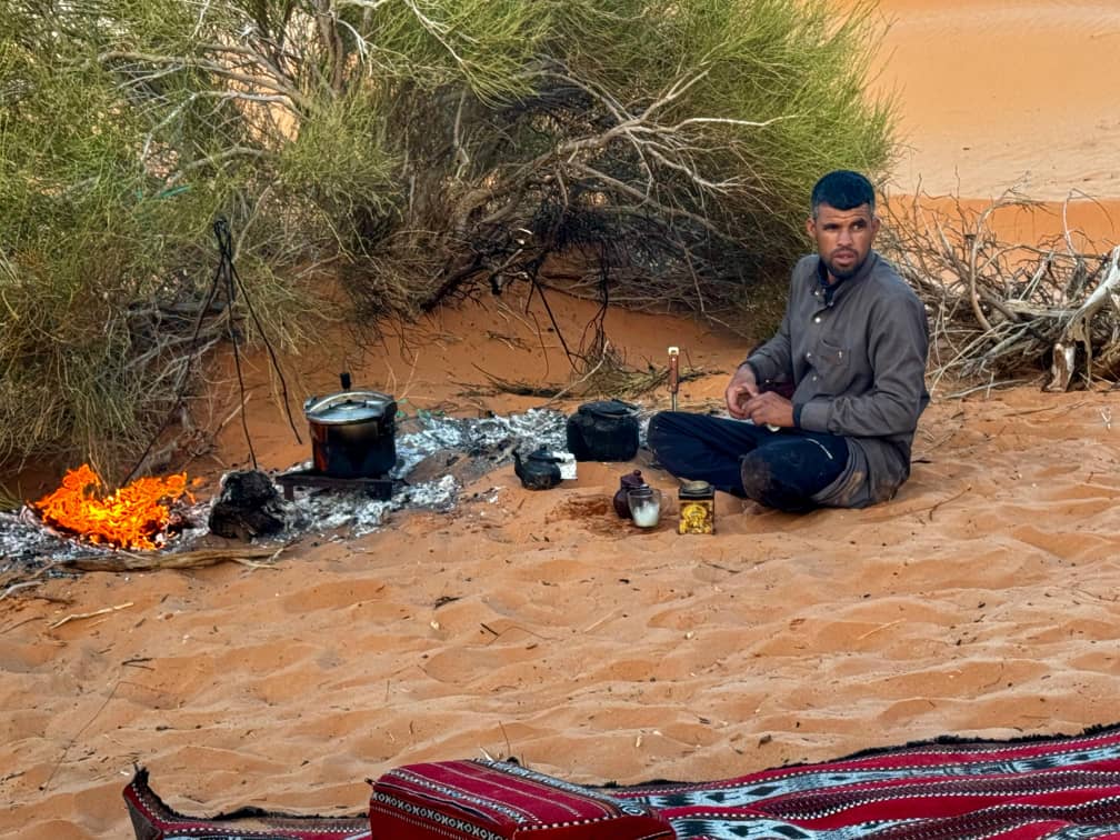 A Saharan guide brewing tea over an open fire in the sand, kettle and pots beside a patterned rug